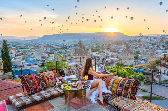Frau sitzt auf einer Hotelterrasse mit Blick &uuml;ber die Landschaft von Kappadokien und bestaunt die aufsteigenden Hei&szlig;luftballons