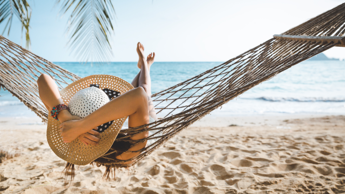 Frau mit Sonnenhut liegt entspannt in einer Hängematte am Strand mit Blick auf das Meer